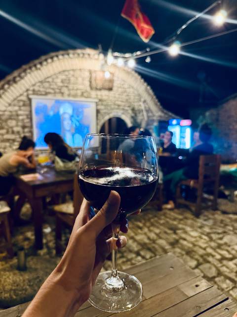 Close-up of a hand holding a glass of red wine inside a cozy stone-walled tavern with friends chatting in the background