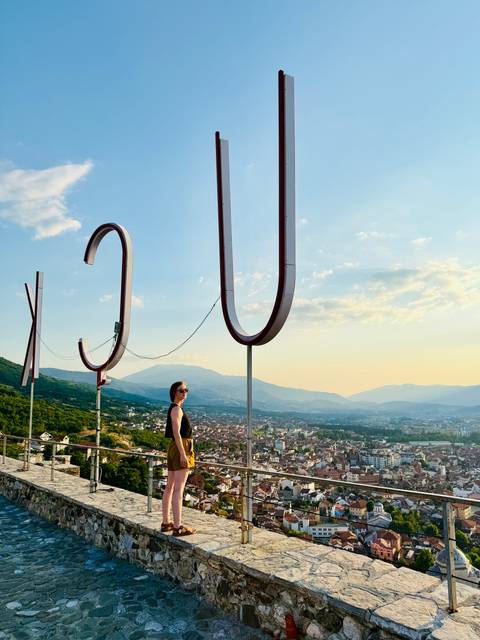 Traveler standing beneath oversized metal letters on a hilltop, city and mountains stretching into the hazy sunset