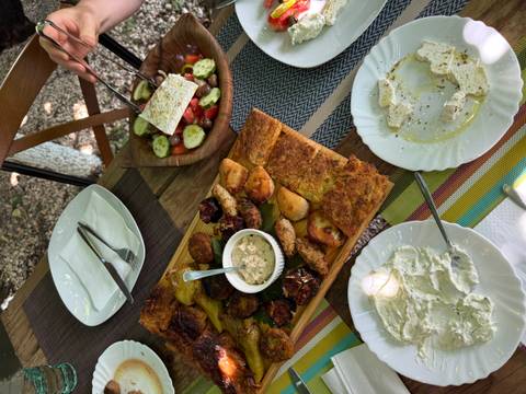 Outdoor table laden with Albanian dishes—grilled vegetables, dips, feta salad and byrek—set under leafy trees