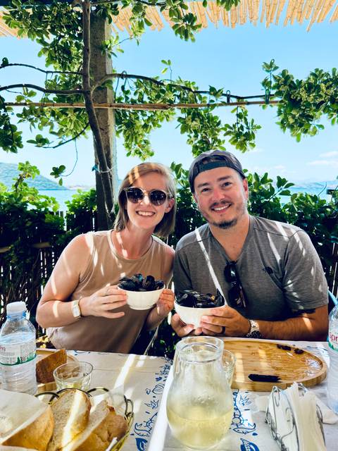 Smiling couple in sunglasses holding bowls of fresh mussels at a shaded waterfront restaurant