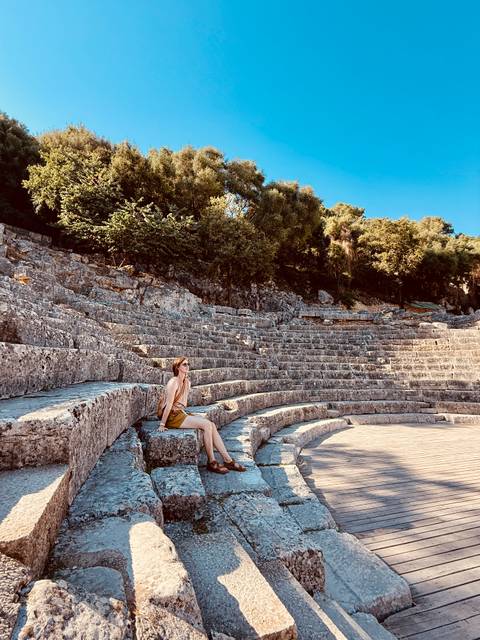 Woman resting on ancient stone tiers of a sun-drenched amphitheater surrounded by trees