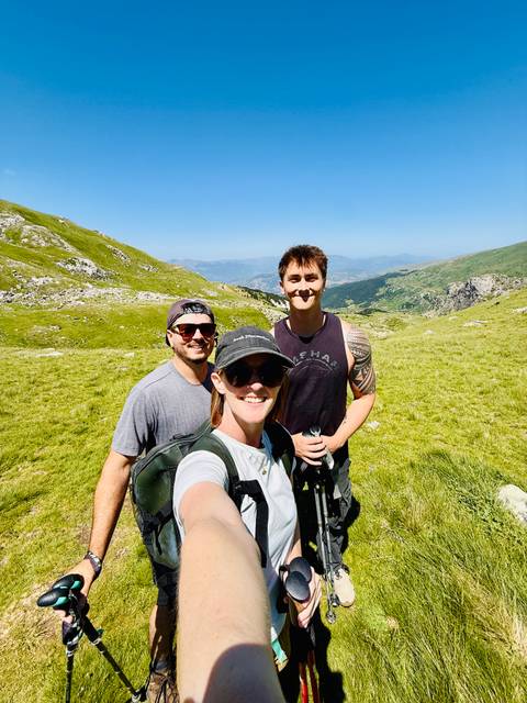 Selfie of three hikers atop a bright green alpine meadow with rolling mountains behind them
