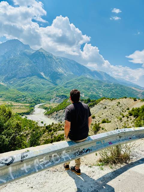 Man gazing over a winding river valley toward towering green mountains under scattered clouds