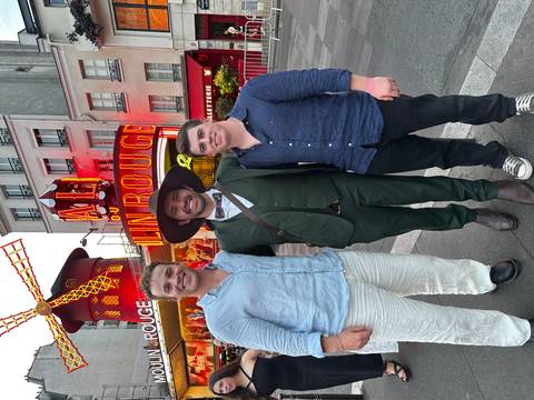 Three friends pose in front of the illuminated Moulin Rouge cabaret in Paris at dusk.