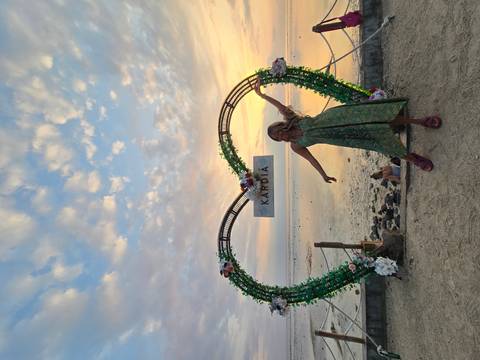 Woman poses beneath a heart-shaped floral arch by the sea during colourful sunset.