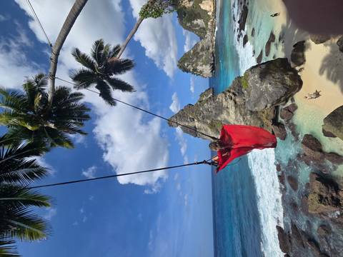 A woman in a red dress soars on a swing above turquoise water and jagged limestone islets.