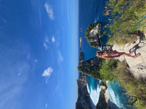 Woman stands on rocky cliff edge with multiple small islands and deep blue sea behind.