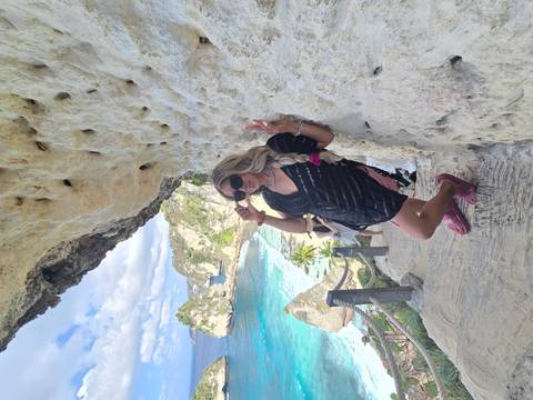 A traveler in sunglasses flashes peace signs while standing on stone steps carved into a dramatic cliff overlooking bright turquoise sea.