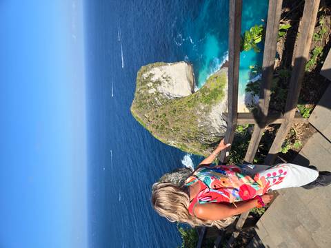 A woman gazes at the famous Kelingking cliff formation rising from deep-blue waters on a sunny day.