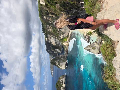 A woman stands on a cliff lookout admiring turquoise waves crashing against white limestone formations.