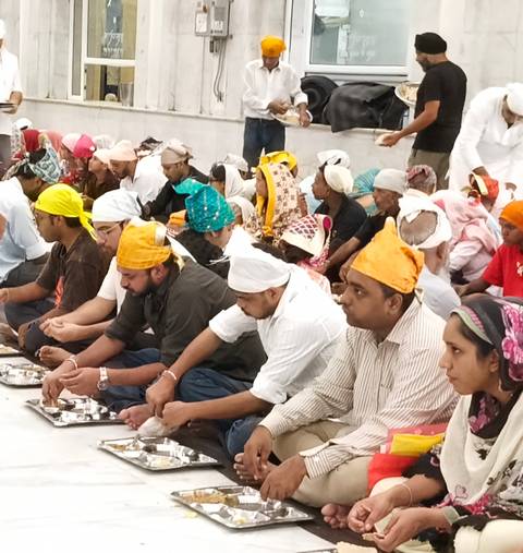 Devotees wearing colorful headscarves sit cross-legged on the floor sharing a communal meal in a temple hall.