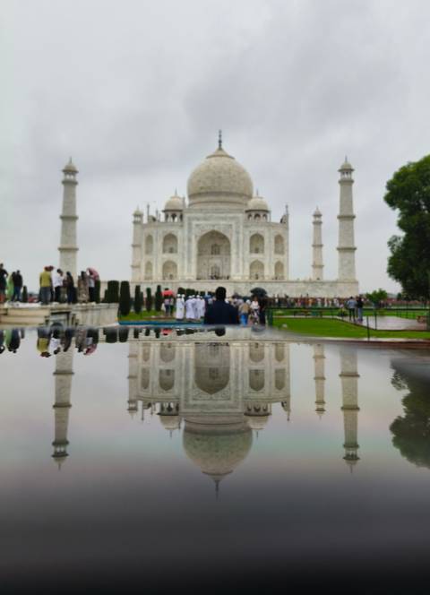 The Taj Mahal reflected in a wet surface on a cloudy day with visitors walking in front.