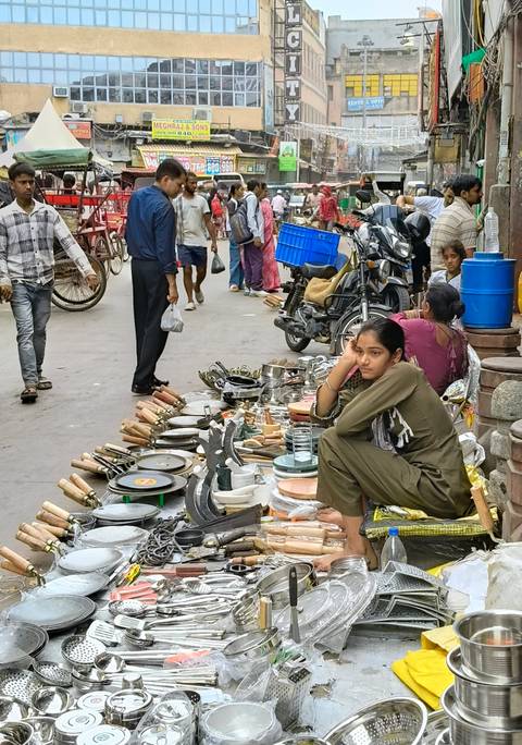 A young street vendor sits among stacks of kitchenware on a busy city lane, looking thoughtful.