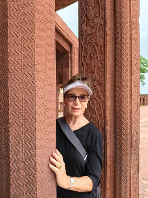 An older woman peeks playfully from behind an ornately carved red sandstone column.