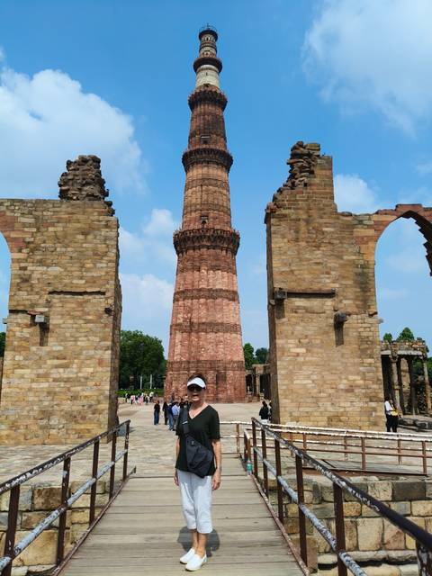 A traveler wearing a visor stands before the soaring stone tower of Qutub Minar framed by ancient arches.