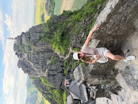 Traveler rests on stone wall with dramatic Mua Cave viewpoint rising behind her.