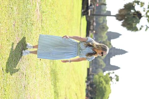 Woman in striped dress walks across a grassy field with Angkor Wat towers behind her.
