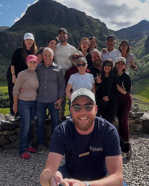 Cheerful tour group poses together against rolling green Scottish hills.