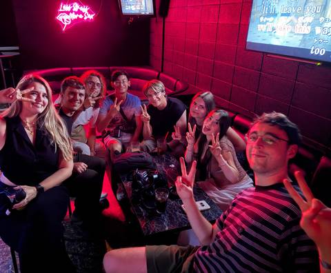 Group of young travellers sitting in a red-lit karaoke lounge flashing peace signs at the camera.