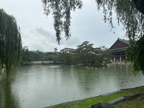 Serene palace pavilion and trees reflected on a rain-dappled pond under overcast sky.