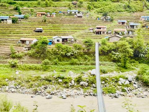 Terraced green hills dotted with small houses above a rocky river in rural Nepal.