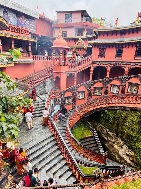 People ascending ornate red spiral stairways adorned with deity statues at a Kathmandu temple complex.