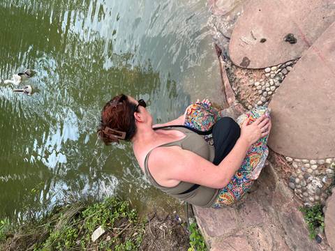 Woman kneels on stones beside a pond reaching toward the water where ducks swim.