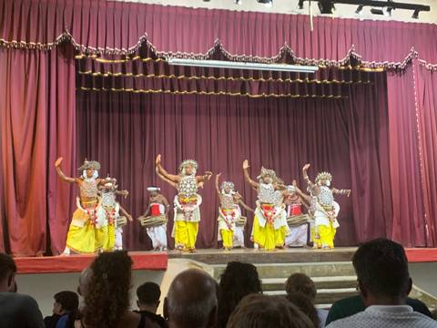 Traditional Sri Lankan dancers in ornate costumes performing on a theatre stage with drummers behind.