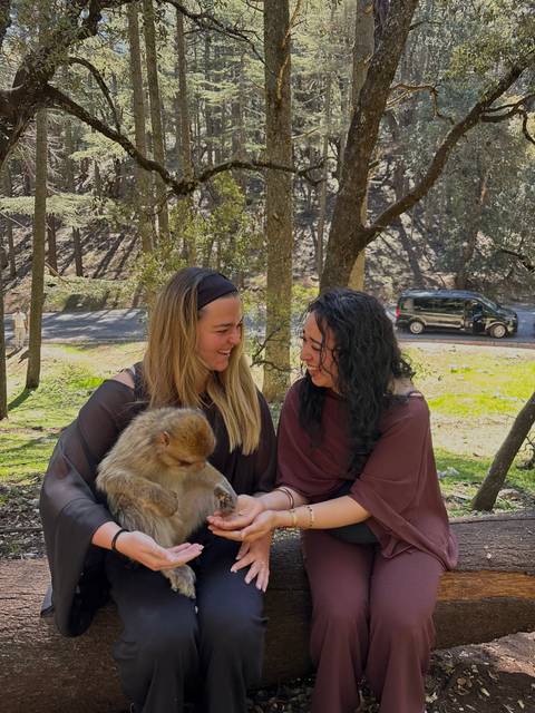 Two friends smiling while holding a young Barbary macaque in a forested roadside area.