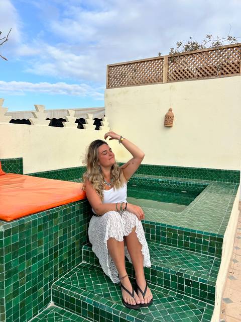 Woman relaxing beside tiled plunge pool on a riad rooftop under blue sky.