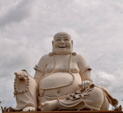 Large laughing Buddha statue with prayer beads against a cloudy sky.