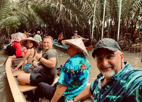 Tourists in a narrow wooden boat wearing conical hats and life jackets on a palm-lined canal.