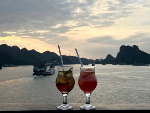 Two cocktails held against a sunset backdrop of karst islands and cruise boats in Ha Long Bay.