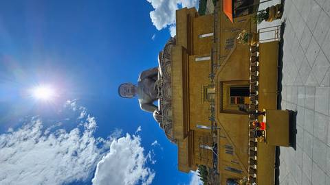 Front-facing view of the giant Buddha statue below a bright sunburst in a clear blue sky, with golden railings and offerings in the foreground.