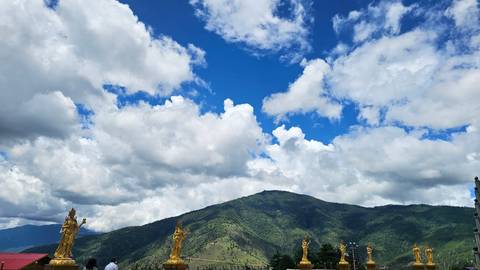 Wide mountain landscape under bright blue sky with dramatic clouds and a row of golden Buddhist statues in the foreground