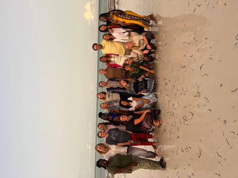 Travel group poses together on a sandy beach with gentle waves at dusk