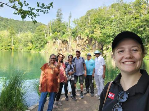 A smiling tour group poses by the emerald lake and limestone cliffs of Plitvice Lakes National Park.