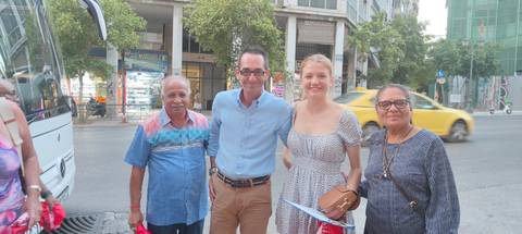 Travellers stand together on a busy city street beside their tour bus, taxis and storefronts behind them.