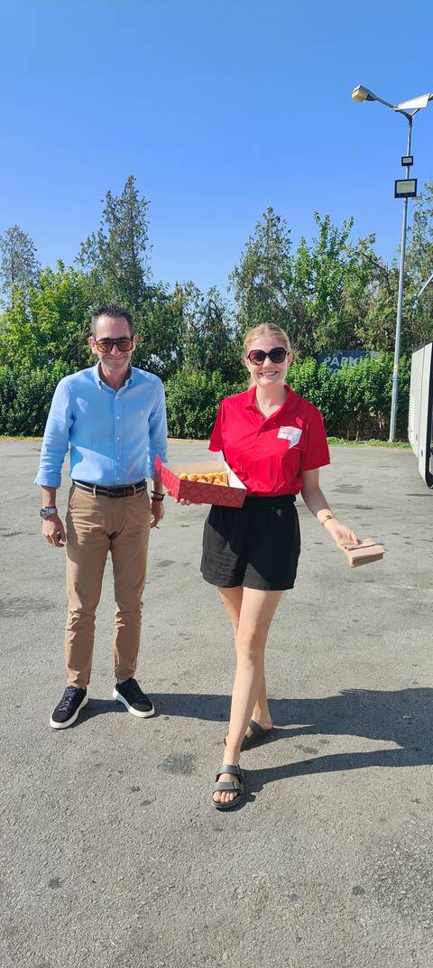 A man and a smiling tour staff member stand on a paved lot near a coach; the staffer holds a box of bite-sized pastries.