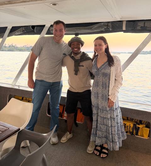 Three travellers smile with their local guide on a covered boat cruising a wide African river at sunset.
