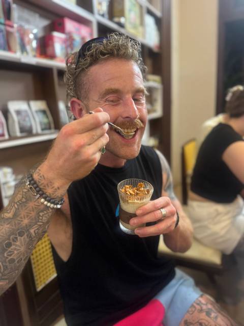 Tattooed traveler enjoys spooning a layered dessert from a glass in a Vietnamese cafe.
