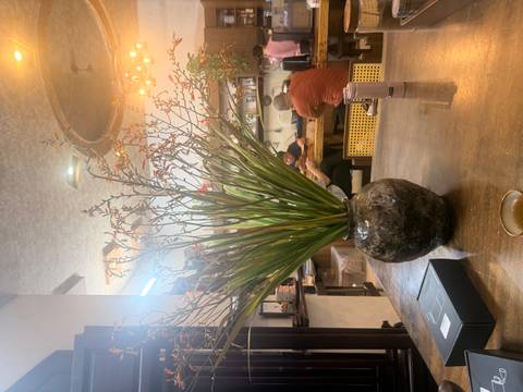 Indoor cafe table with a large vase of foliage; patrons converse in the softly lit background.