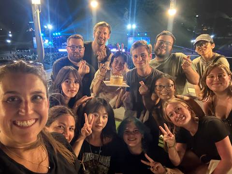 Large group selfie of smiling travelers holding a small cake at an outdoor night event.