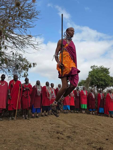 Maasai warriors in red shukas perform a high jumping dance while others watch in a semicircle.