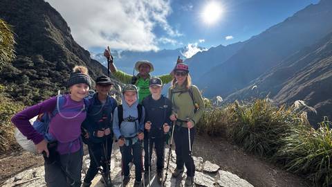A family of hikers smiling on the Inca Trail with dramatic Andean peaks and bright sun overhead.