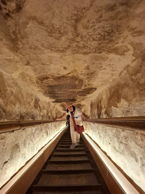 Visitor climbs steep wooden ramp inside the narrow descending passage of a pyramid.