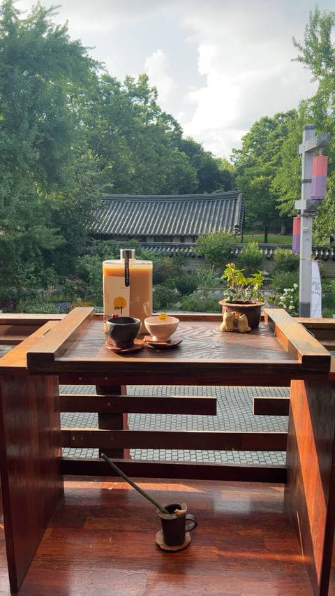 A wooden tray holds traditional drinks and ceramics on a balcony overlooking a Korean courtyard garden.
