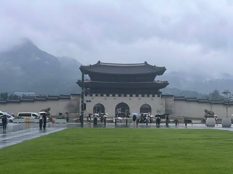 Gwanghwamun Gate stands against misty mountains and rain as visitors with umbrellas cross the plaza.