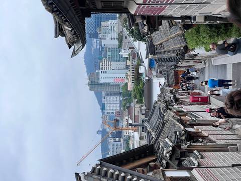 Visitors walk down the traditional tiled-roof alley of Bukchon Hanok Village toward Seoul's modern skyline.