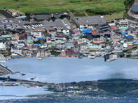An elevated view shows a coastal town and tranquil blue bay surrounded by hills.
